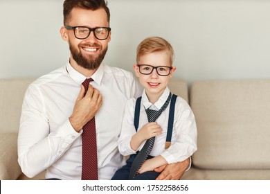 A happy bearded businessman in a strict shirt tie and glasses sits with his young son in a similar outfit on the sofa and looks at the camera with a smile - Shutterstock ID 1752258656