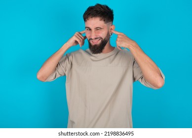 Happy Bearded Caucasian Man Wearing Casual T-shirt Over Blue Background Ignores Loud Music And Plugs Ears With Fingers Asks To Turn Off Sound