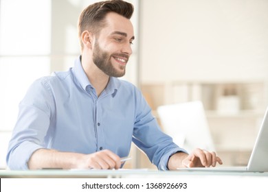 Happy Bearded Employee In Blue Shirt Sitting In Front Of Laptop While Surfing In The Net For Some Particular Information