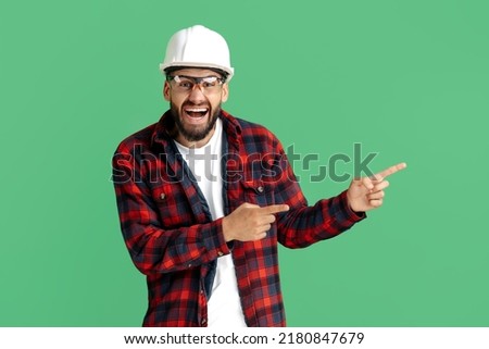 Happy bearded engineer or constructor man in casual outfit pointing finger away over green background.
