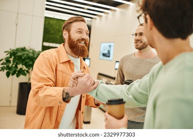 happy bearded entrepreneur shaking hands with business partner near smiling colleague while holding paper cups during coffee break in lobby of modern office space, success and cooperation concept - Powered by Shutterstock