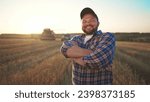 Happy bearded farmer in cap looking at camera standing on agricultural wheat field with combine harvester and dump truck on background. Harvesting, farming at sunset. Food production, agribusiness.