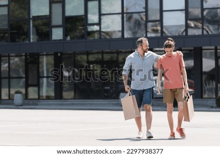 happy bearded father hugging teenage son while walking from mall with shopping bags