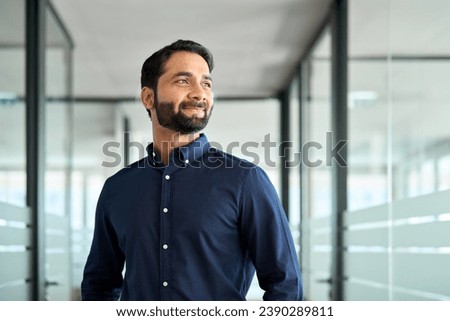 Happy bearded Indian business man leader looking away standing in office hallway. Professional smiling businessman manager executive or male employee or entrepreneur thinking of future success.