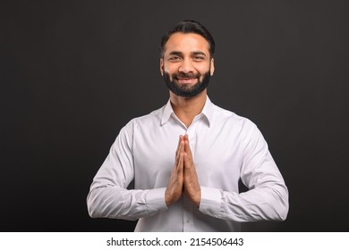 Happy bearded Indian man in formal white shirt greeting or thanking you with traditional gesture namaste standing isolated on black background. Male office employee welcoming, holding arms in pray