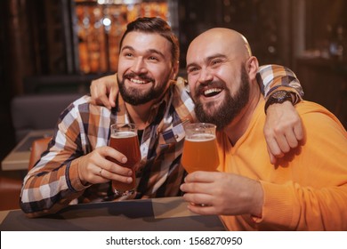 Happy Bearded Man Enjoying Drinking Beer With His Best Friend. Male Friends Hugging, Holding Up Their Beer Glasses, Celebrating At The Bar