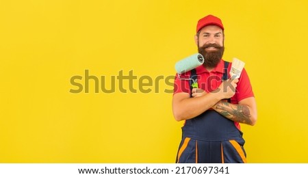 happy bearded man housepainter in work clothes hold paint roller and brush on yellow background