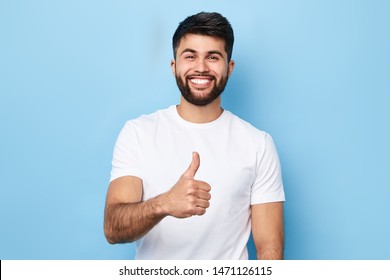 Happy Bearded Man In Stylish White T-shirt With Beaming Smile Showing Thumb Up, Isolated Over Blue Background, Studio Shot. Good Job, Well Done. Agreement Concept