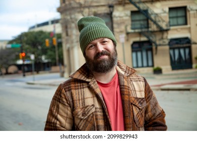 A happy bearded man wearing a green knitted beanie and a brown plaid coat smiles for a portrait in a downtown setting on a sunny day.
