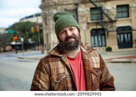 A happy bearded man wearing a green knitted beanie and a brown plaid coat smiles for a portrait in a downtown setting on a sunny day. Stock foto © 