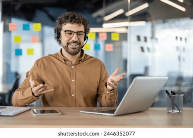 A happy bearded man wearing headphones talks cheerfully in a video conference in a modern office setting. - Powered by Shutterstock