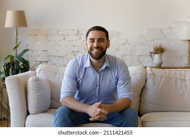 Happy Bearded Millennial Guy Making Video Call From Home, Sitting On Couch, Looking At Webcam With Toothy Smile. Business Man, Teacher, Couch Chatting Online On Virtual Conference. Headshot Portrait