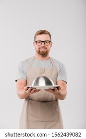 Happy Bearded Waiter In Apron And Eyeglasses Passing You Cloche With Cooked Food While Standing In Front Of Camera In Isolation