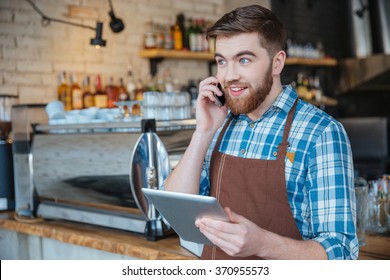 Happy bearded young waiter talking on cell phone and using tablet in cafe - Powered by Shutterstock