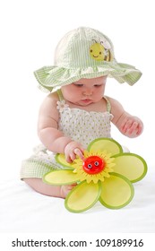 Happy Beautiful 6 Month Old Baby Girl In Green Seersucker Sun Hat And Sun Dress Sits And Plays With A Big Yellow Toy Daisy. Pastels, Isolated On White Background, Vertical, Copy Space.