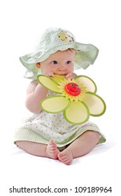 Happy Beautiful 6 Month Old Baby Girl In Green Seersucker Sun Hat And Sun Dress Sits And Plays With A Big Yellow Toy Daisy. Pastels, Isolated On White Background, Vertical, Copy Space.