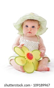 Happy Beautiful 6 Month Old Baby Girl In Green Seersucker Sun Hat And Sun Dress Sits And Plays With A Big Yellow Toy Daisy. Pastels, Isolated On White Background, Vertical, Copy Space.