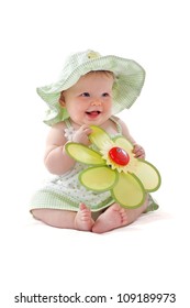 Happy Beautiful 6 Month Old Baby Girl In Green Seersucker Sun Hat And Sun Dress Sits And Plays With A Big Yellow Toy Daisy. Pastels, Isolated On White Background, Vertical, Copy Space.