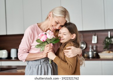 Happy Beautiful Affectionate Middle Aged Mom Receiving Spring Flowers Bouquet Hugging Cute Teen Child Kid Daughter Standing At Home In Kitchen Enjoying Surprise Moment Celebrating Mothers Day Holiday.