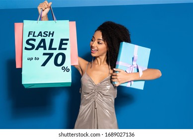 Happy Beautiful African American Woman In Silver Dress Holding Gift Box And Looking At Shopping Bags On Blue Background, Flash Sale Illustration