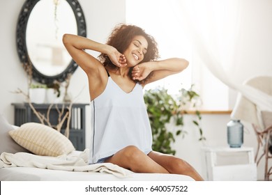 Happy beautiful african girl in sleepwear stretching smiling with closed eyes sitting on bed at home woke up in the morning on a sunny day. - Powered by Shutterstock