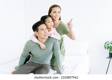 Happy Beautiful Asian Family Sitting On Bed And Looking Up Together Through The Window In Bedroom At Home. Young Korean Mother Point Up With Finger. Emotional Happy Family Enjoying Tender Moment.