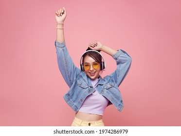 Happy beautiful Asian teen woman in yellow glasses listening music in headphones and dance on pink background. 