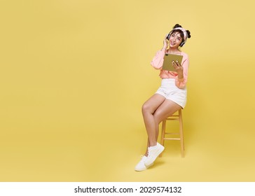 Happy beautiful Asian teen woman sitting on chair and hand holding tablet with wearing wireless headphones listening to music on yellow copy space background.  - Powered by Shutterstock