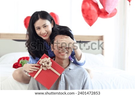 Happy beautiful Asian woman covering her boyfriend eyes, giving present gift box while sitting on white bed in bedroom, romantic lover couple celebrate memorable anniversary on Valentine Day together.