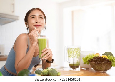 A happy and beautiful Asian woman is enjoying her healthy green smoothie in the kitchen after her morning workout. Healthy lifestyle, diet, wellbeing, detox drinks - Powered by Shutterstock