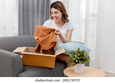 Happy Beautiful Asian Woman Opening Box With Ordered Clothes At Home On Couch. Online Shopper Customer Smiling And Holding Clothes. Delivery Service Concept.