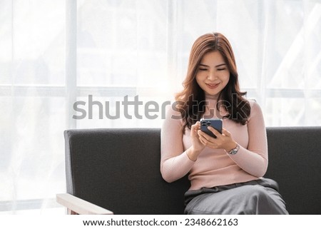 Happy beautiful Asian woman sit on sofa hand holding mobile phone chatting with friends and playing social media. Young female looking at smartphone cellphone browsing internet at home living room..