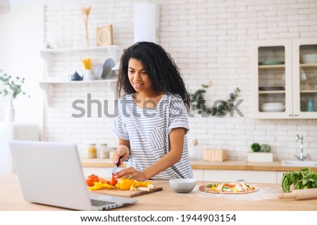 Happy beautiful biracial woman enjoying cooking and filming it, looking for a recipe on the laptop in the modern kitchen, food blogger concept, healthy lifestyle, online learning cooking class concept