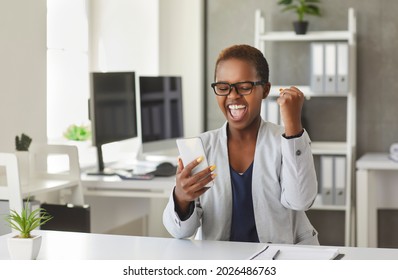 Happy Beautiful Black Woman Sitting At Office Desk, Holding Smartphone, Fist Pumping And Smiling. Young Business Lady Is So Glad And Excited To Get Deal Approval Email On Mobile Phone. Success Concept