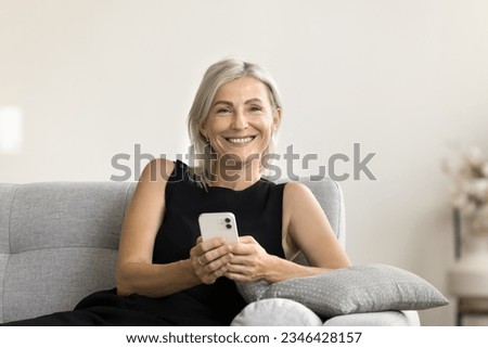 Happy beautiful blonde mature woman in black holding white cellphone, resting on home sofa with gadget, looking at camera with toothy, promoting online media service, application, domestic Internet