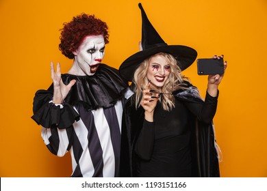 Happy Beautiful Blonde Witch And Scary Clown In Halloween Costumes Making Selfie On Smartphone Isolated Over Orange