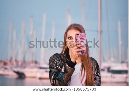 Happy beautiful blonde woman taking photos with a smartphone in a sea port. Young smiling girl using mobile phone. Sea and boats on background.
