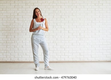 Happy beautiful brunette dressed in strip overalls, standing and holding red paper heart, white brick wall on background