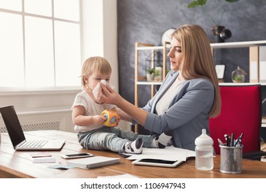 Happy Beautiful Business Mom Wiping Child's Nose While Working With Documents In Office. Business, Motherhood, Multitasking And Family Concept.