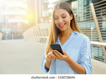 Happy beautiful business woman typing on her smart phone while standing in modern city street. Copy space.