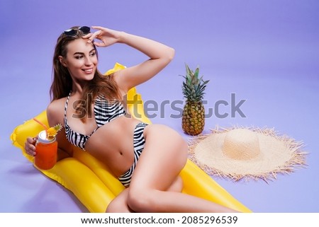 Happy beautiful caucasian adult girl wears in bikini lyes on inflatable yellow matress while holds cocktail on purple background . Summer concept. Travel concept. Relax concept 