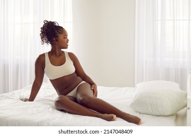 Happy Beautiful Chubby Young Black Woman In Underwear Sitting On Bed At Home And Looking Away. Relaxed African American Plus Size Female Model In Soft Bra And Underpants Posing In Bedroom Interior