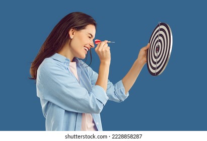 Happy beautiful confident young brunette woman in casual shirt squints eye and aims dart arrow at shooting target in her hand. Side profile view studio shot. Setting goals, strategy, success concept - Shutterstock ID 2228858287