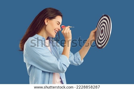 Happy beautiful confident young brunette woman in casual shirt squints eye and aims dart arrow at shooting target in her hand. Side profile view studio shot. Setting goals, strategy, success concept