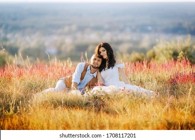 Happy Beautiful Couple In Love Are Sitting And Hugging In Flowers On A Field On A Hill. Stylish Wedding Photo Session In Rustic Style. Romantic Portrait Of The Bride And Groom On Their Wedding Day