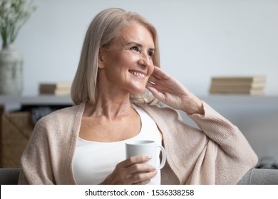 Happy beautiful dreamy old senior middle aged woman sit on sofa relax at home hold cup drink coffee tea looking away thinking dreaming enjoy stress free peaceful mood wellbeing alone in living room