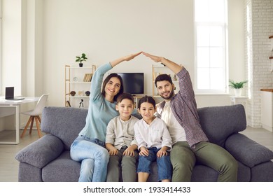 Happy Beautiful Family Of Four Sitting On Sofa At Home. Smiling Mom And Dad Doing Arm Roof Gesture Above Little Son And Daughter. Mortgage, House Insurance, Children Protection, Future Plans Concept