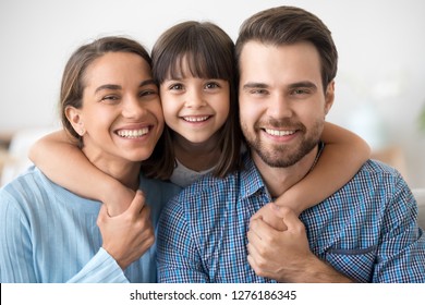 Happy Beautiful Family Of Three Dad Mom And Little Cute Daughter Portrait, Child Girl Smiling Embracing Mother And Father Posing With Parents Looking At Camera, Kid Hugging Mommy Daddy Headshot