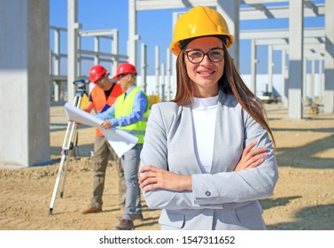Happy beautiful female architect on construction site. She is smiling and satisfied with her job, behind her construction engineers planning and talking about the project, teamwork
