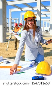 Happy beautiful female architect on construction site. She is smiling and satisfied with her job, behind her construction engineers planning and talking about the project, teamwork